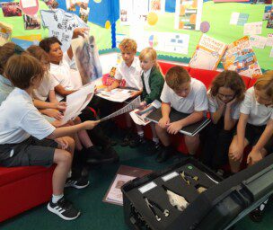 A group of school children are sitting on red sofas with colourful display boards behind them. They're all looking at A3 size pieces of paper and looking into a large plastic box containing a skull and lower jaws of animals.