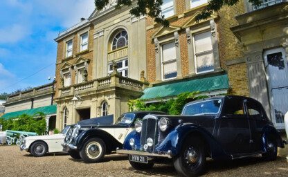 Three classic cars parked on a gravel driveway outside a large regency house on a bright sunny day.