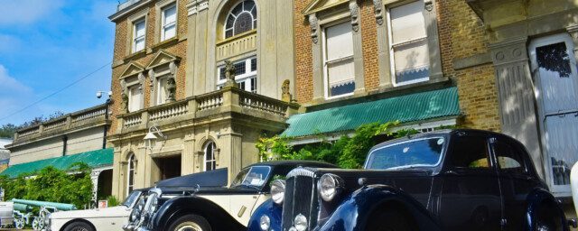 Three classic cars parked on a gravel driveway outside a large regency house on a bright sunny day.