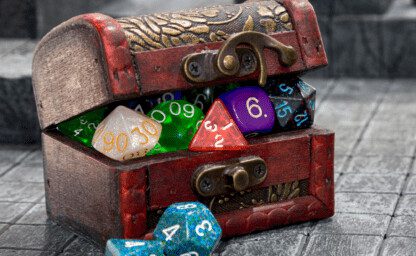 A wooden looking treasure chest with gold decorative in lay on the lid and front. The lid is rounded has a latch on the front. The box is slightly held open by all the different shapes and colourful dice that are inside. Two blue dice are on the floor to one side of the box.