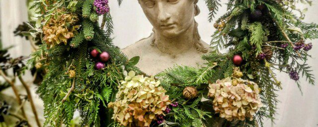 A circular wreath with evergreen foliage dried hydrangea flower heads and purple berries, hanging from a traditional bust of a female on a wooden plinth.