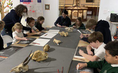 A large table with school children sitting all the way around it. They are all drawing onto black paper using white chalk with a white female with brown curly hair standing behind one of the children. There is a row of various skulls positioned down the middle of the table.