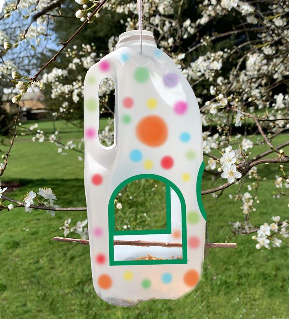A photograph of a plastic milk bottle hanging in a tree with blossom on the branches and green grass on the ground. The milk bottle has multi coloured spots all over it and some arch shaped holes cut in towards the bottom with green colour round the arches. The arches are big enough to allow for birds to fly in. There is a twig pushed through the bottom, level with the bottom of the arch sharped holes, as a perch for birds and some seed can just be seen at the bottom of the milk bottle.