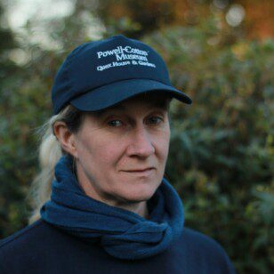 Photograph of a white female with fair skin tone and blonde hair in a pony tail. Wearing a dark blue baseball cap, with white writing on the front saying, Powell-Cotton Museum Quex House & Gardens. A slightly lighter blue scarf and dark blue sweatshirt with white writing across the front to one side saying, Powell-Cotton Museum Quex House & Gardens. Blurred green foliage is in the back round.