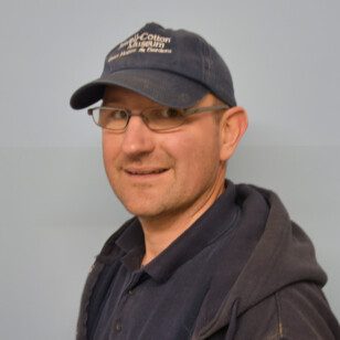 Photograph of a white male positioned in a slightly sideways pose. The male has brown eyes and rectangular glasses with a little stubble and is smiling. He is wearing a dark blue cap with White writing on the front saying Powell-Cotton Museum Quex house and Gardens. A dark blue polo style t-shirt and an unzipped, dark blue hooded jumper.