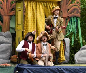 Three white adults in fancy dress. Two are playing instruments and all have their mouths open as if they're singing. They are outside with trees in the background and stage scenery is behind the adults.