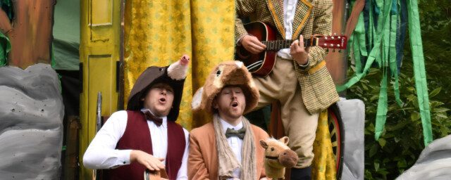 Three white adults in fancy dress. Two are playing instruments and all have their mouths open as if they're singing. They are outside with trees in the background and stage scenery is behind the adults.